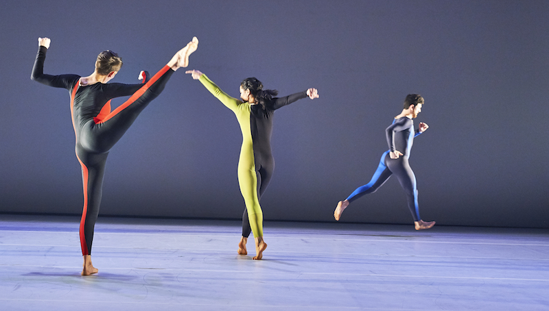 Three dancers in unitards. One lifts her leg to the side and another crosses her legs as her feet are in relve. Both of their backs are to the audience. Another dancer in the background runs to the right.